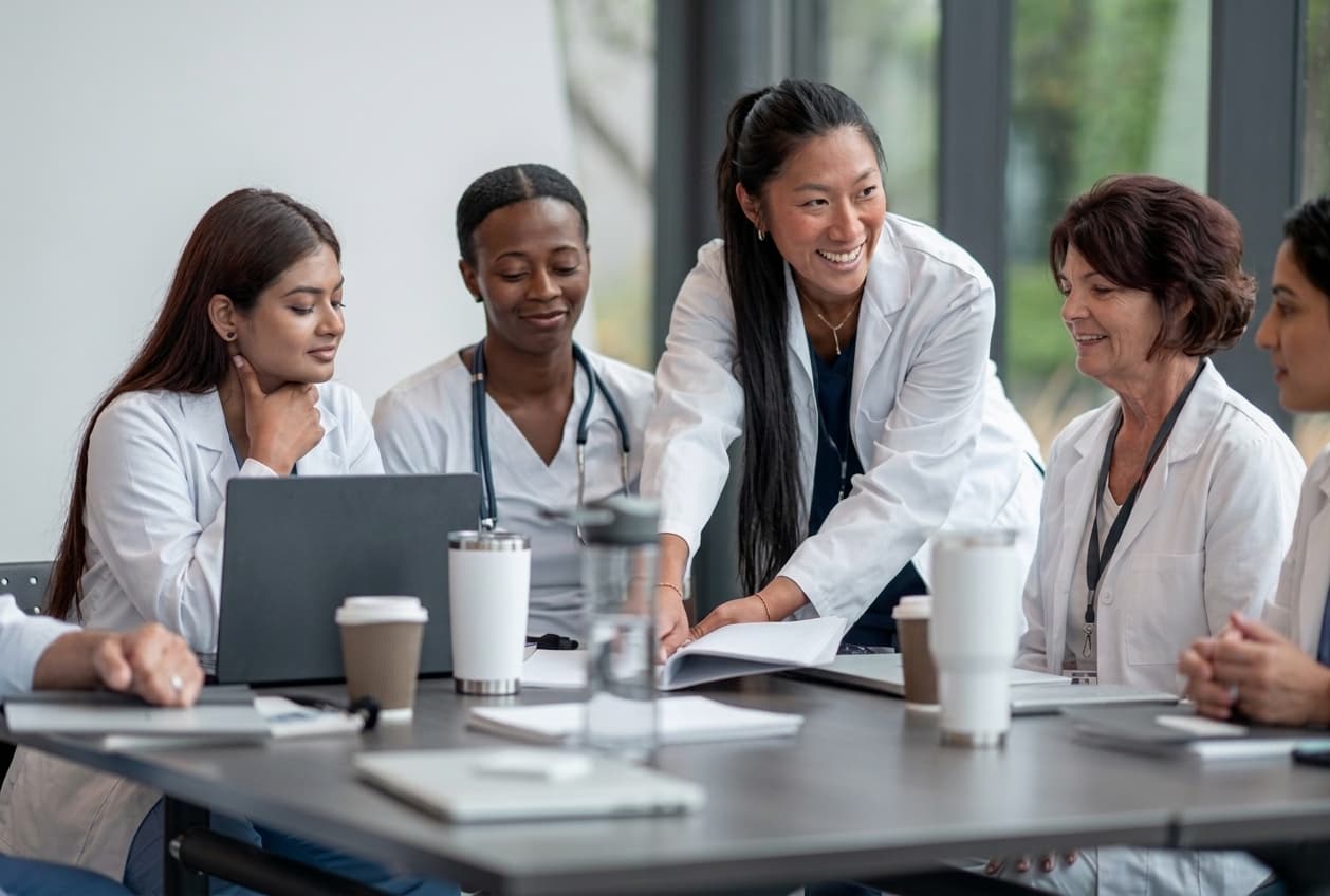 Team of women healthcare providers in white coats reviewing marketing performance reports during a leadership meeting