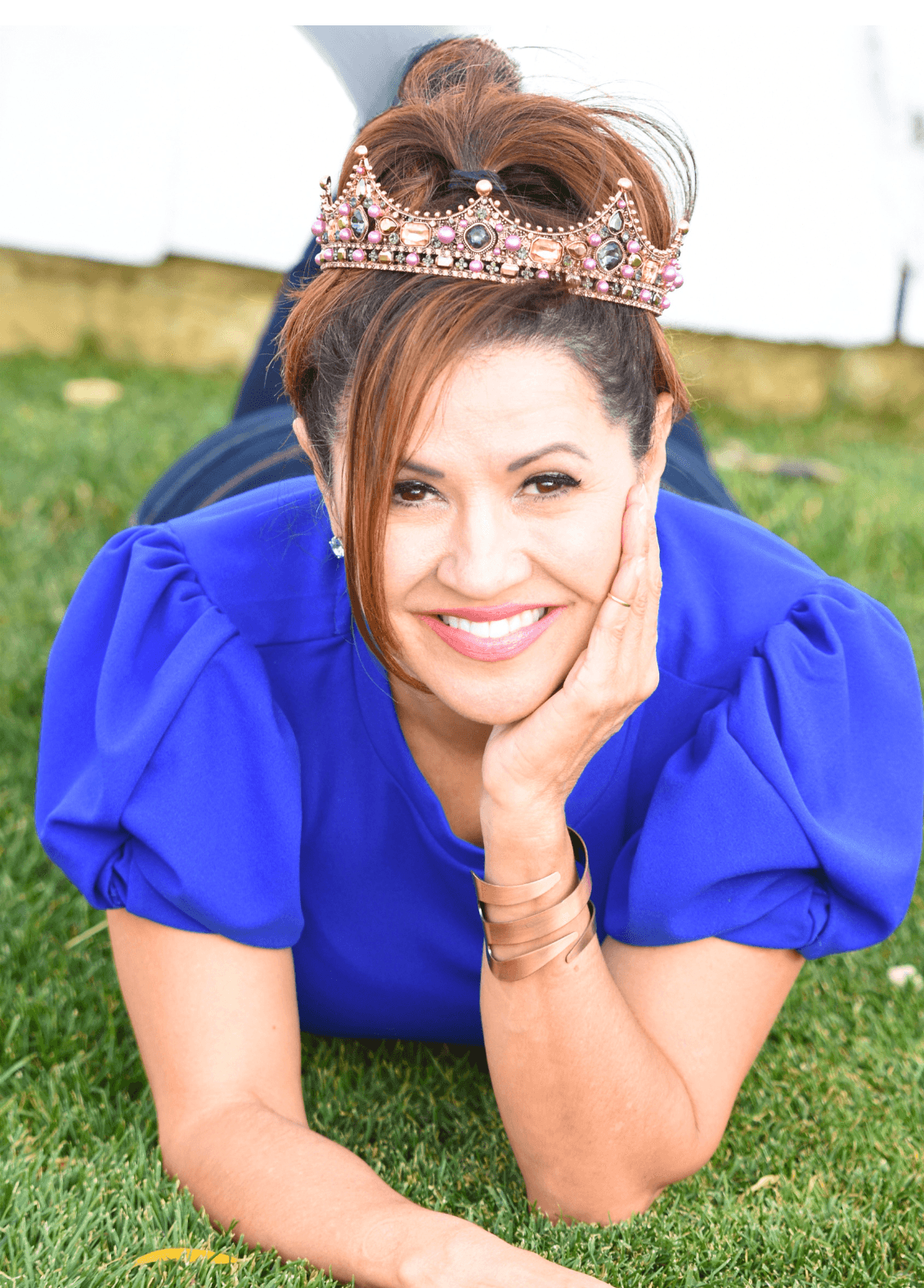 Kris Mitchell in her playful Queen Kristiana author persona, wearing a tiara, lying on the grass
